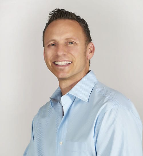 Headshot of a man in a light blue dress shirt, smiling in front of a white background