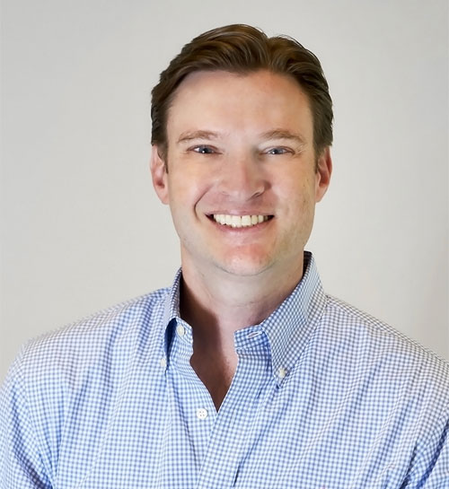 Headshot of a man in a light blue shirt, smiling in front of a grey background